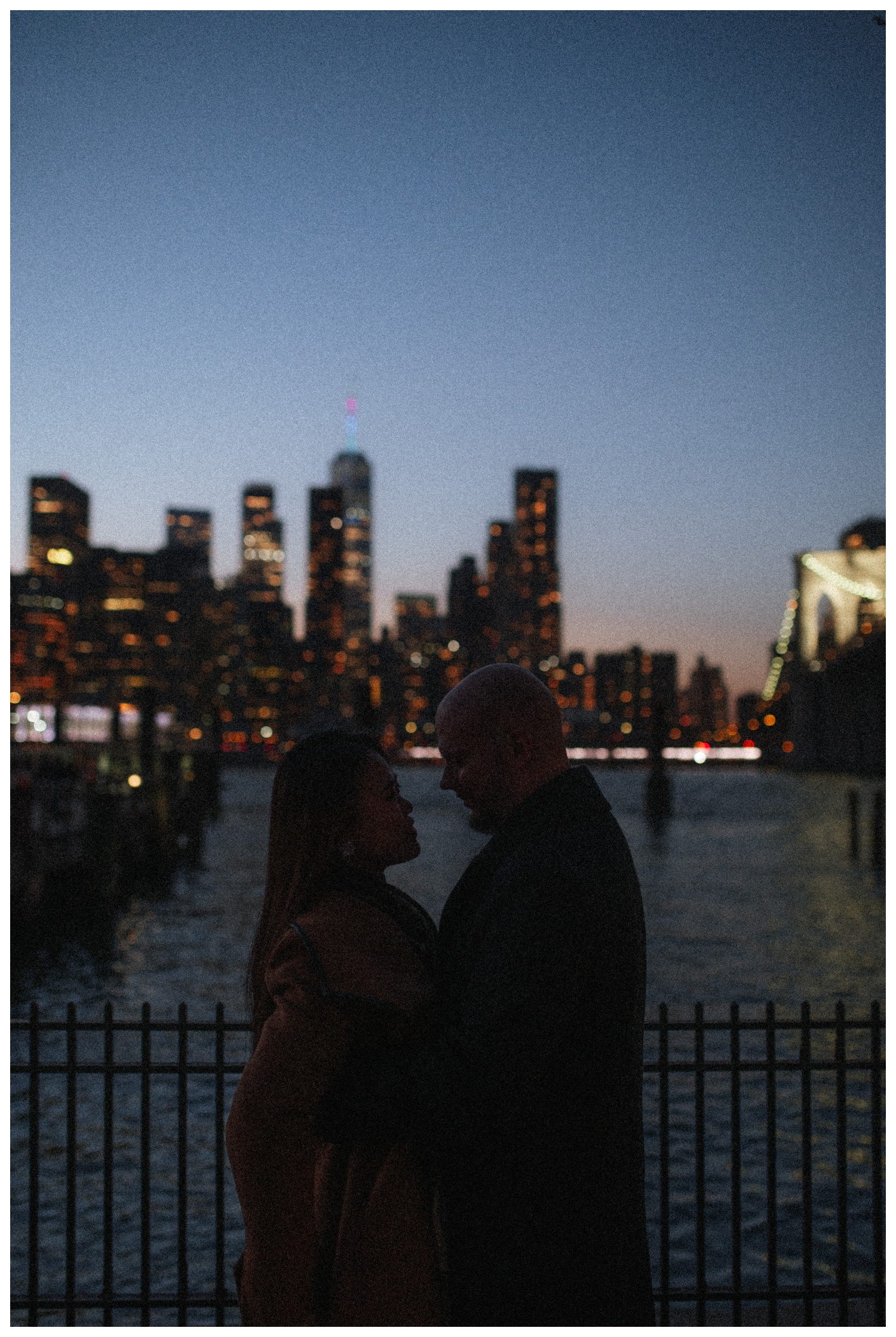 Engaged couple standing close in front of the Manhattan skyline while in Brooklyn. New York City wedding photographer. Brooklyn wedding photographer. Parr Photo Co.